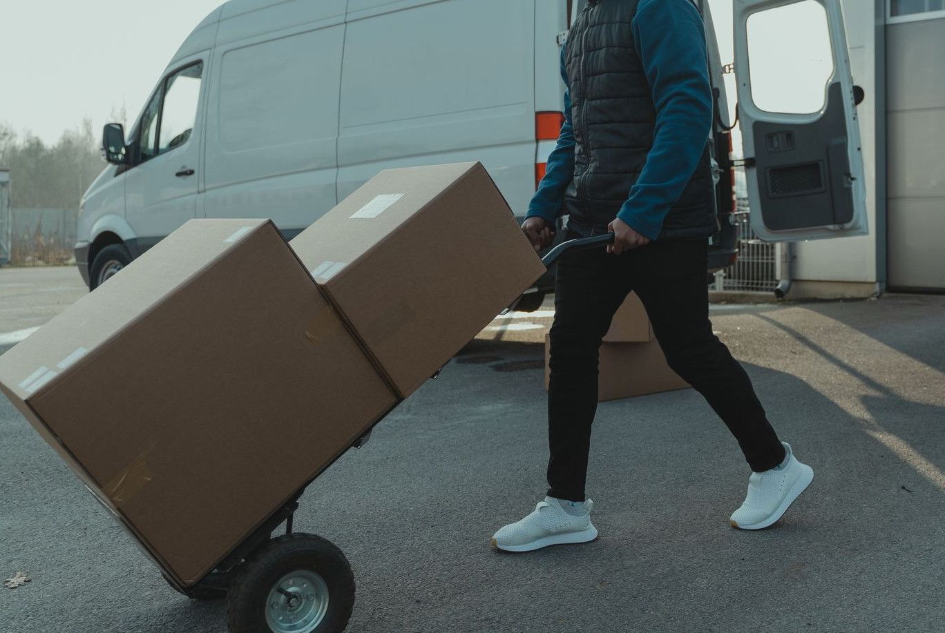 Courier pushing a trolley with boxes near a van at a warehouse, showcasing delivery services.